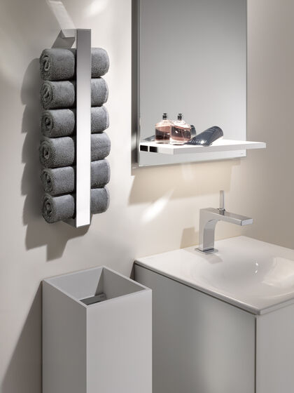 Waschbecken Edition 11 von KEUCO, das an einer weißen Wand installiert ist. Darüber hängt ein Spiegel. Links daneben befindet sich ein Mülleimer, darüber befindet sich ein Handtuchhalter mit gerollten Handtüchern.