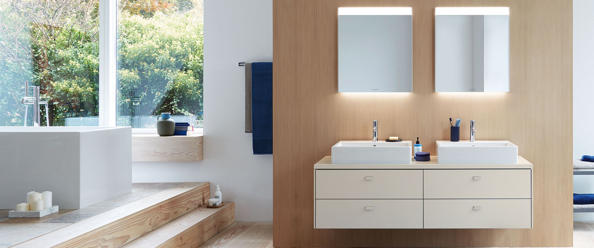 Badambiente mit Doppelwaschbecken und helle Badmöbel- Beleuchtete spiegel und eine Badewanne.