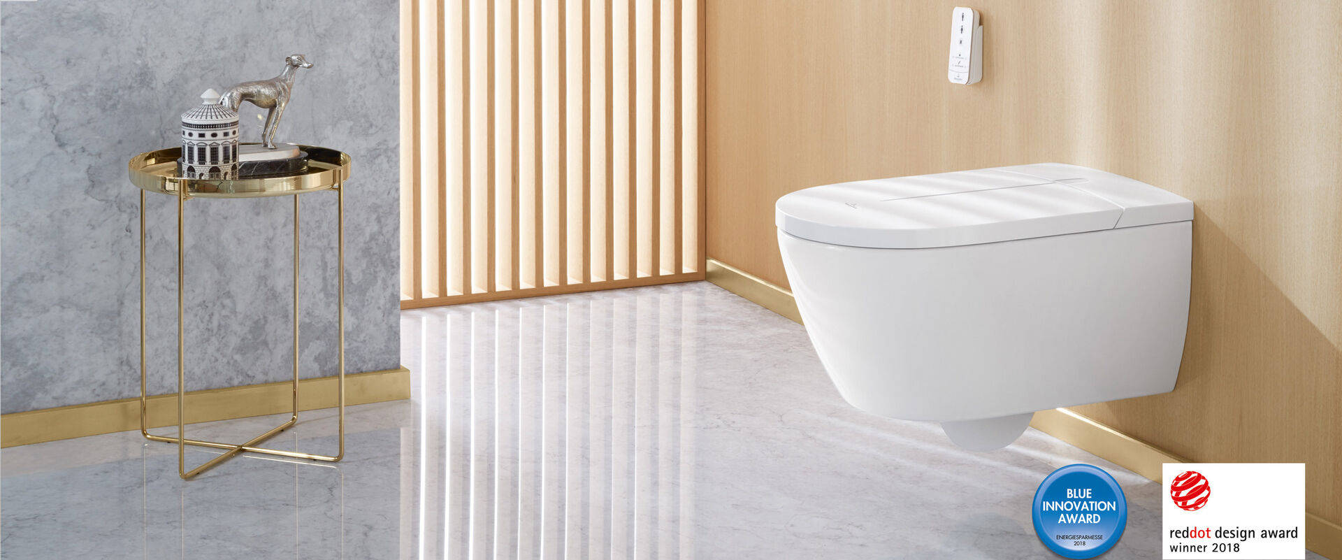 Dusch-WC Szene mit goldfarbenem Beistelltisch, Lamellenfenster und dem innovativen Dusch-WC ViClean-I 100 von Villeroy & Boch. Logos im Bild verweisen auf Design-Awards.