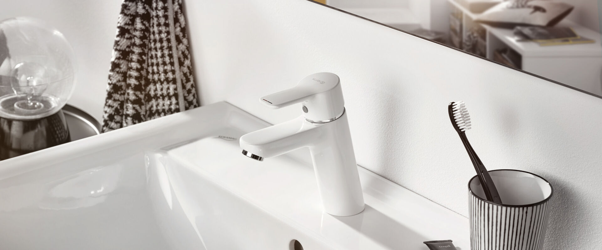 Weißer Wasserhahn Pure Funktion von Kludi. Installiert an einem weißen Waschbecken, auf dessen Rand ein Gefäß mit Zahnbürste steht.