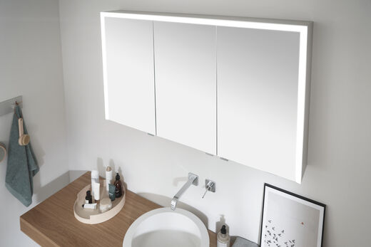 Ansicht von oben auf den rechteckigen Spiegelschrank Elegant-Line 2.0 von SPRINZ mit angeschaltetem, dreiseitigem LED-Licht und drei Türen, in der Korpusfarbe Aluminium. 