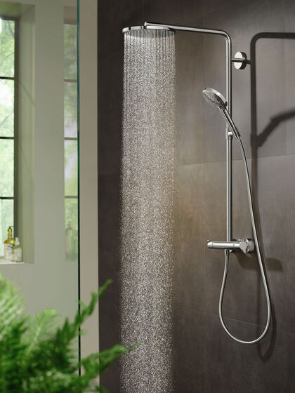 Raindance Select Duschsystem von hansgrohe, dass an einer grau gefliesten Wand installiert ist. Aus der Kopfbrause strömt Wasser. Im linken Teil des Bildes ist im Hintergrund ein Fenster zu erkennen, auf dem Fensterbrett steht Duschgel. Im Vordergrund links befindet sich eine grüne Pflanze.