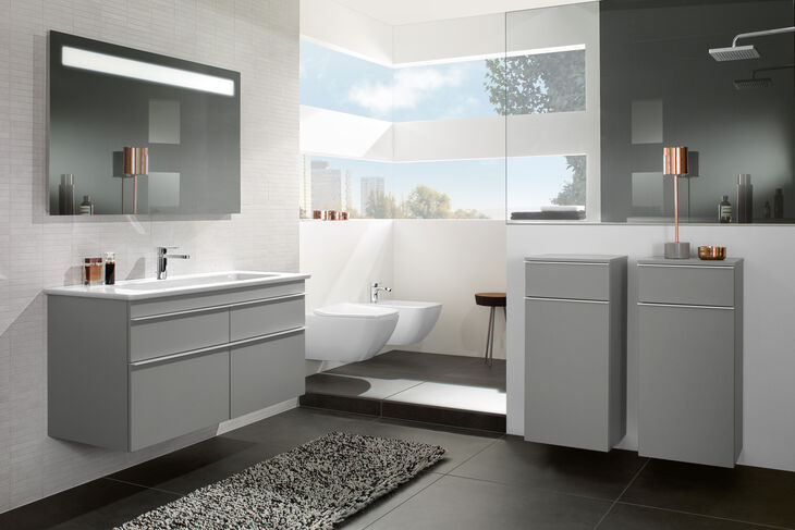 Modernes Badezimmer mit grauen Badmöbeln und Chromleisten. Wandmontierte Badmöbel und LED Lichtspiegel. Toilette und Bidet im Hintergrund.