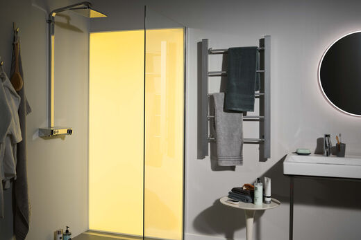 Gelb beleuchtete Duschwand Premium LED von Spritz. Sie befindet sich in einem ansonsten grau gehaltenem Badezimmer. Links von der Dusche ist ein Handtuchhalter, darunter ein Tisch als Ablagemöglichkeit sowie ein Waschbecken mit Spiegel zu erkennen.