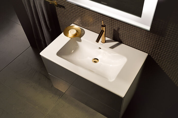 Elegantes Waschbecken und goldener Badarmatur und mittigem goldenen Ablauf. Badmöbel in Braun und beleuchtetr Spiegel.