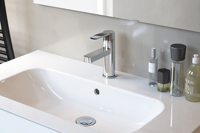 Geo Wasserhahn von Ideal Standard, der an einem weißen Waschbecken installiert sind. Auf dem Beckenrand stehen Hygieneartikel. Im Hintergrund ist ein Teil eines Spiegels zu erkennen.