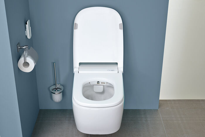 Blick in ein geöffnetes Dusch-WC von VitrA vor einer blauen Wand montiert. Das V-care Modell ist spülrandlos und bietet zahlreiche Komfortfunktionen.