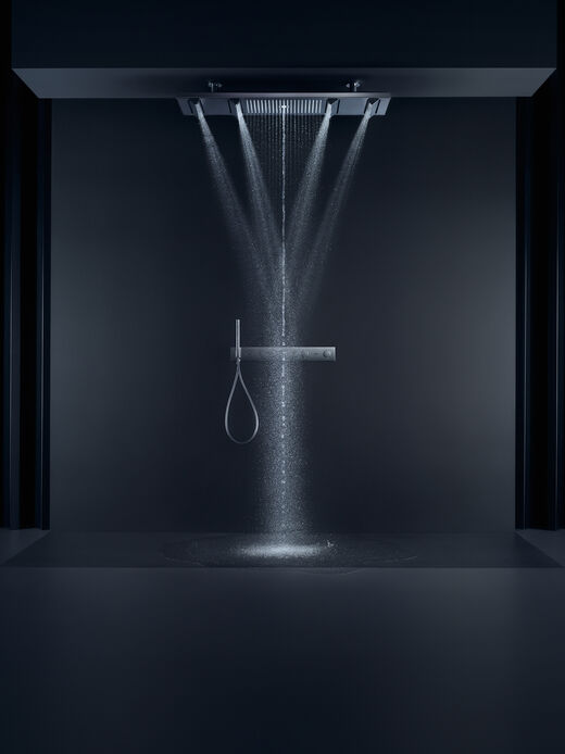 Dusche in einem schwarz gehaltenem Raum, in dem eine Shower Heaven Regendusche installiert ist. An einer rechteckigen, länglichen Kopfbrause sind mehrere zusätzliche Brausen quer über die eigentliche Brause installiert, aus denen in unterschiedliche Richtungen Wasser fließt.