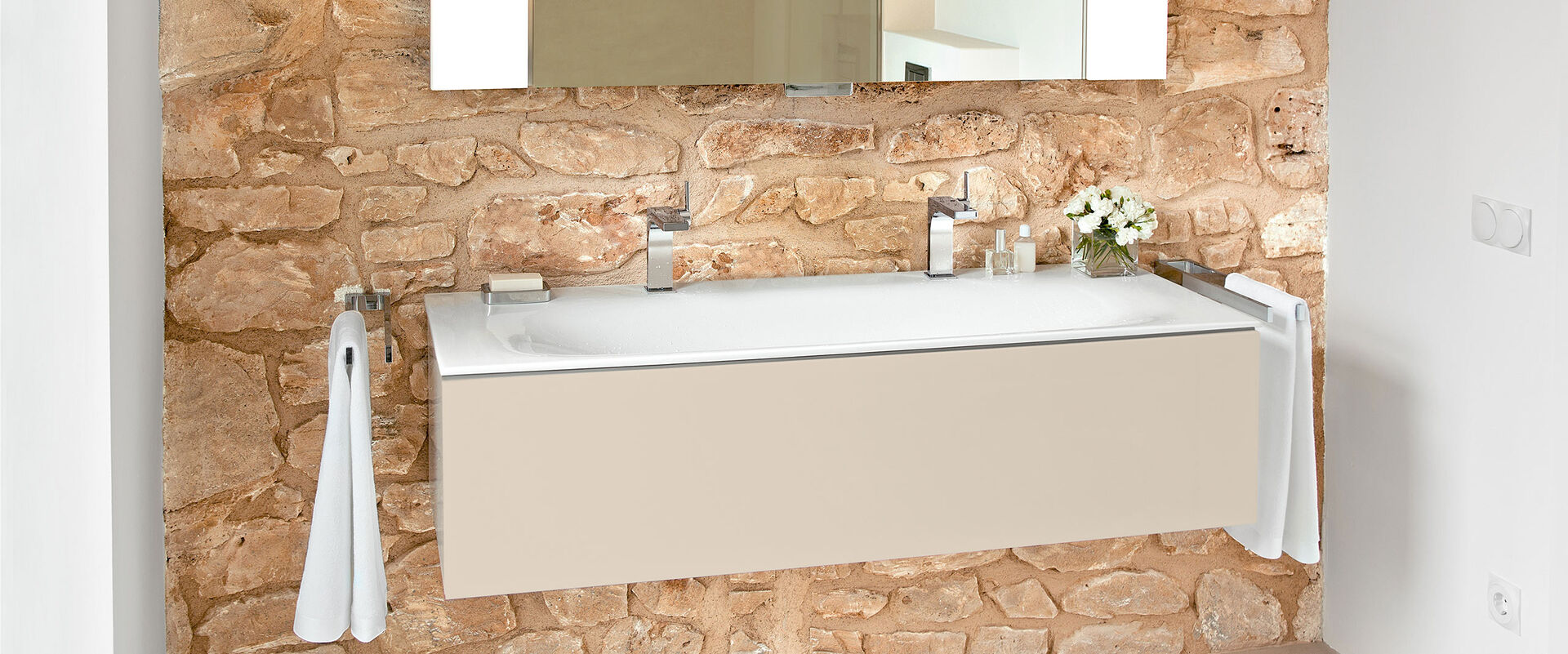 Waschbecken Edition 11 von Keuco, das an einer rustikal gemauerten Wand installiert ist.  Auf dem Waschbecken stehen Hygieneartikel, daneben hängen Handtücher, darüber hängt ein Spiegel.