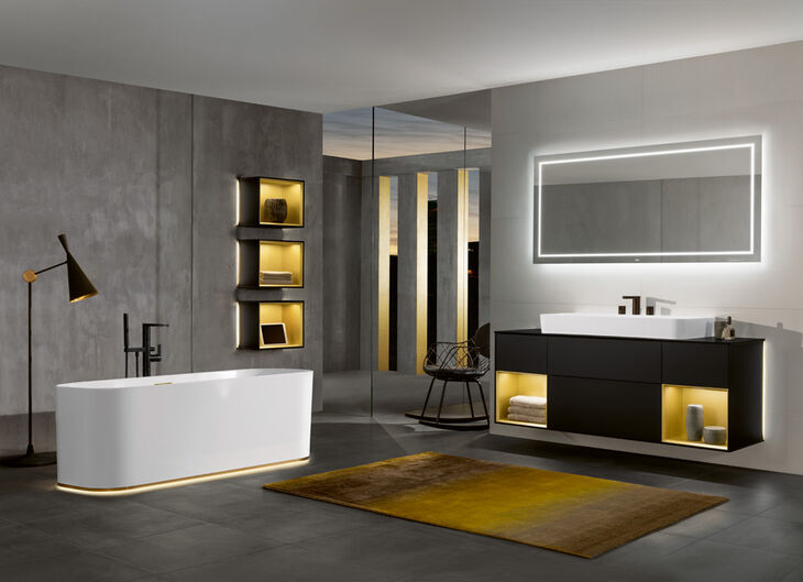 Badezimmer von Villeroy und Boch mit grauen Wänden und Fußböden. Links steht eine weiße Badewanne, Rechts befindet sich ein Waschbecken mit Unterschrank und Spiegel. Im ganzen Raum sind goldene Farbakzente gesetzt.