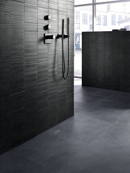 In Grau gehalten Raum, in dem eine mit schmalen Fliesen geflieste Wand steht. An dieser Wand ist eine Duscharmatur befestigt, am Boden ist eine Geberit Clean Linke Duschrinne eingebaut.