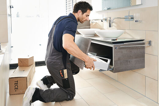 Ein Fachhandwerker baut einen Waschplatz ein und trägt dabei Schutzschuhe. Er kniet vor dem Badmöbel und schiebt den Auszug ein.