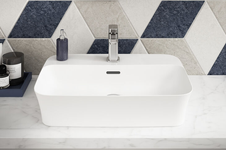 Ideal Standard Ipalyss Weißes Waschbecken Ipalyss von Ideal Standard, das sich auf einem Waschtisch befindet. Darüber ist ein Wasserhahn installiert. Die Wand hinter dem Waschbecken ist rautenförmig gefliest.