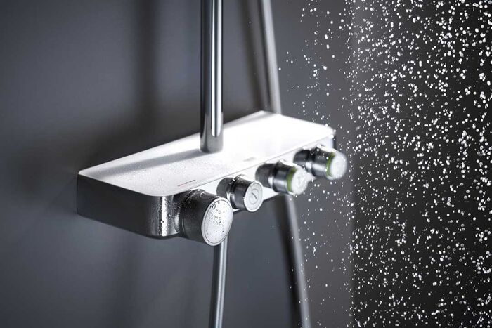 GROHE Smartcontrol Duscharmatur, die an einer grauen Duschwand installiert ist. Wassertropfen sind rechts im Bild zu erkennen. Es sind insgesamt vier verschiedene Betätigungsknöpfe mit unterschiedlichen Funktionen angebracht. Eine Hand betätigt den mittleren Betätigungsknopf auf der vorderen Seite.