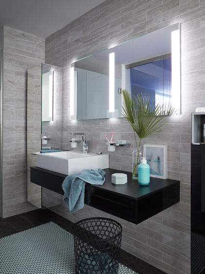 Waschbereich in einem Badezimmer mit Blumenvase und einem extrabreiten Lichtspiegel, der in der Mitte noch einen Beleuchtungsstreifen aufweist.
