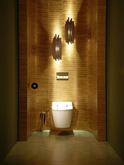 Premium-Gästebad von Duravit in Nachtaufnahme: Vor einer goldfarbenen Wand thront das SensoWash Dusch-WC, beleuchtet von darüber hängende, eleganten Designer-Lampen.