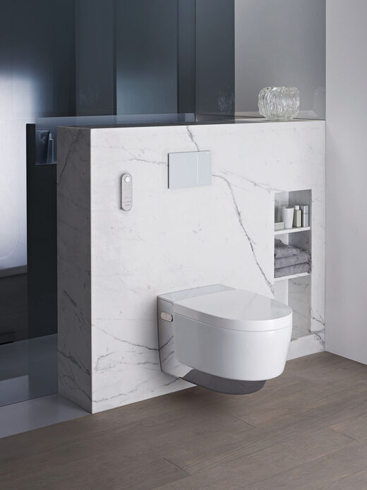 Elegante Dusch-WC-Situation mit dem Modell AquaClean Mera von Geberit an einer weißen Marmor verkleideten Vorwandinstallation mit integrierten Ablagefächern und Fernbedienung.