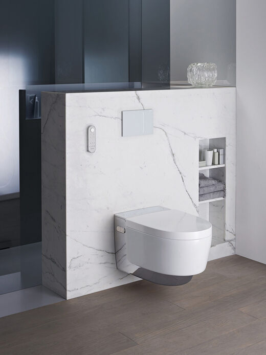 Geberit AquaClean Mera Elegante Dusch-WC-Situation mit dem Modell AquaClean Mera von Geberit an einer weißen Marmor verkleideten Vorwandinstallation mit integrierten Ablagefächern und Fernbedienung.
