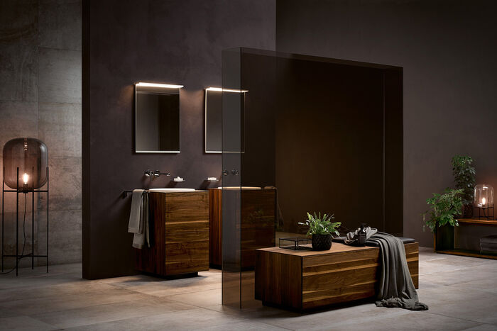In Brauntönen gehaltenes Badezimmer EDITION LIGNATUR von KEUCO. Im Vordergrund befindet sich eine Sitzgelegenheit mit gläsernen Raustrenne, im Hintergrund befinden sich zwei Waschbecken mit Unterschränken und Spiegeln.