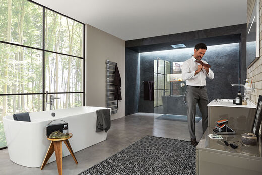 Modernen Mann mit Anzughose bindet sich eine Kravatte und steht in einem großen Badezimmer mit begehbarer Dusche, einer freistehenden Badewanne und einem Waschplatz.