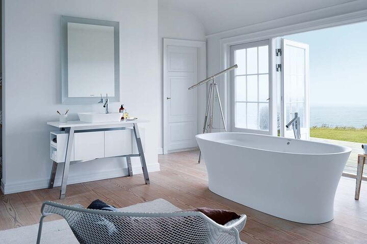 Weisses Badezimmermit einem Waschplatz und Spiegel sowie einer ovalen freistenden Badewanne. Blick auf Dünenlandschaft aus dem Bad.