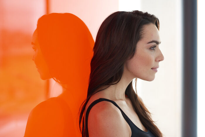 Frau mit langen dunklen haaren lehnt an einer orangenen Wand, in der sie sich spiegelt.
