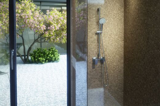 Teil eines Badezimmers, in dem eine Dusche installiert ist, in der die Handbrause Evo von Ideal Standard befestigt ist. Links ist ein Fenster, von dem aus man in den Innenhof mit Baum blickt.