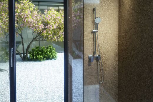 Ideal Standard Evo Teil eines Badezimmers, in dem eine Dusche installiert ist, in der die Handbrause Evo von Ideal Standard befestigt ist. Links ist ein Fenster, von dem aus man in den Innenhof mit Baum blickt.