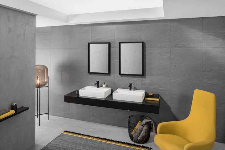 Badezimmer mit grauen Wänden und hellen Decken. An einer Wand ist ein Waschtisch montiert, auf dem sich zwei Villeroy und Boch Memento 2.0 Waschbecken befinden. Darüber hängt ein Spiegel. Rechts befindet sich ein gelber Sessel, links eine Lampe.