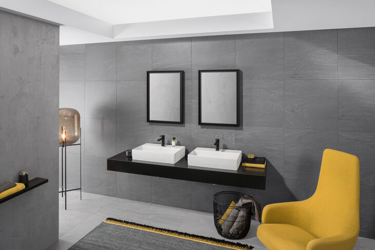 Villeroy & Boch Memento 2.0 Badezimmer mit grauen Wänden und hellen Decken. An einer Wand ist ein Waschtisch montiert, auf dem sich zwei Villeroy und Boch Memento 2.0 Waschbecken befinden. Darüber hängt ein Spiegel. Rechts befindet sich ein gelber Sessel, links eine Lampe.