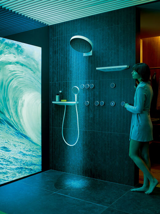 Das Duschsystem RainTunes von hansgrohe kann über RainButtons oder über ein RainPad (Touchscreen) gesteuert werden. Nächtliche Szene mit angeschaltetem Duschzenario, laufender Kopfbrause und einer Frau, die das RainPad bedient.