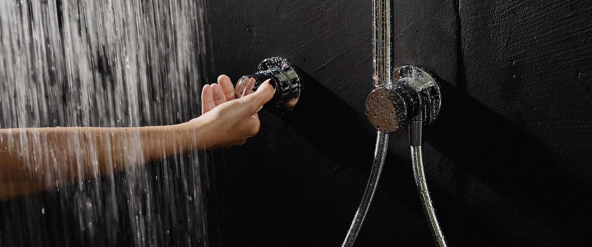 Keuco Ixmo Badarmatur, die an einer robusten schwarzen Wand installiert ist. Die Armatur wird gerade von einer weiblichen Hand betätigt, über die Wasser aus einem nicht sichtbaren Brausekopf läuft.