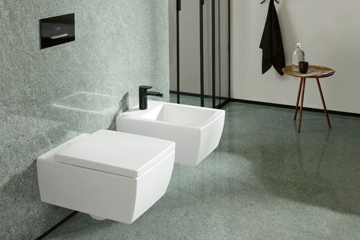 WC- und Bidet-Ensemble vor grünlicher Wand aus der Serie Memento 2.0 von Villeroy & Boch. Eckiges Design mit schwarzen Armaturen kombiniert.