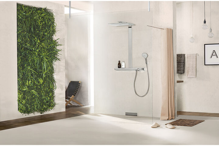 Helles Badezimmer, in dessen Dusche eine hansgrohe rainmaker select Kopfbrause installiert ist. Vor der Dusche liegen Pantoffeln, im Hintergrund hängen Handtücher sowie ein Bild an der Wand. Links befindet sich ein Gang, an dessem Wand grüne Pflanzen wachsen.