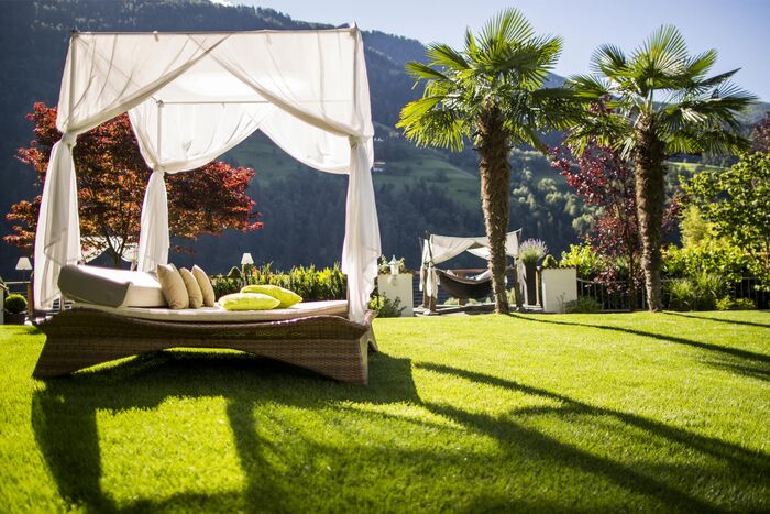 Gartendoppelliege mit weißem Baldachin auf der Wiese des Quellenhofes in Meran mit zwei Palmen daneben.