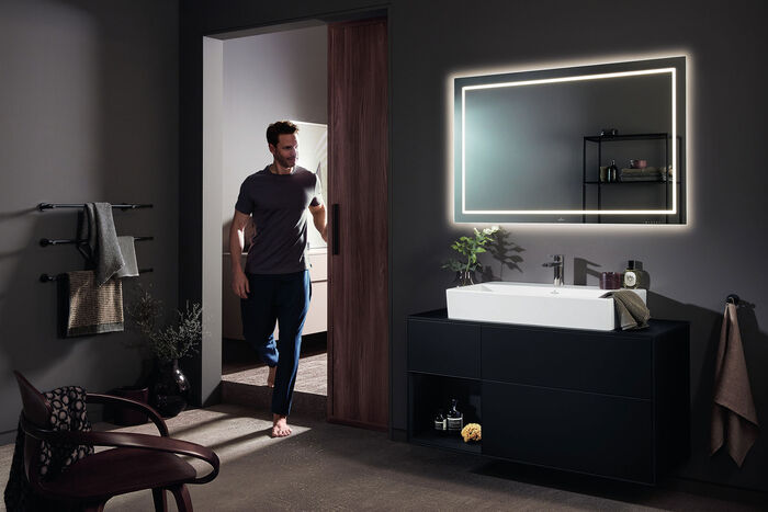 Villeroy & Boch Badezimmerszene bei Nacht, in der ein Mann den Raum betritt und man rechts einen beleuchteten Waschplatz/Spiegelschrank mit einem weißen Memento 2.0 Waschbecken sieht.