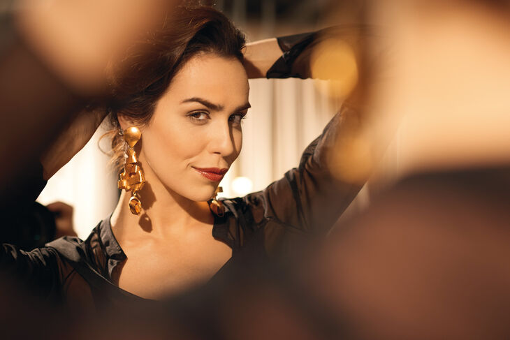 Sinnliche Frau schwarz gekleidet steht vor einem Spiegel. Sie trägt einen großen goldenen Ohrring.
