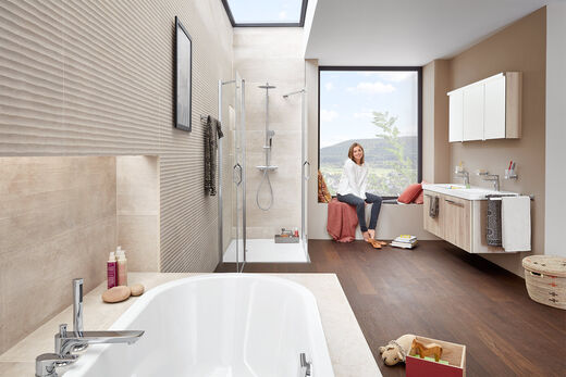 DIANA Bad M Eine junge Frau sitzt am Fenster in Ihrem Badezimmer auf einer Sitzband. Vor ihr seitlich befindet sich der Waschplatz, eine Dusche und eine Badewanne.