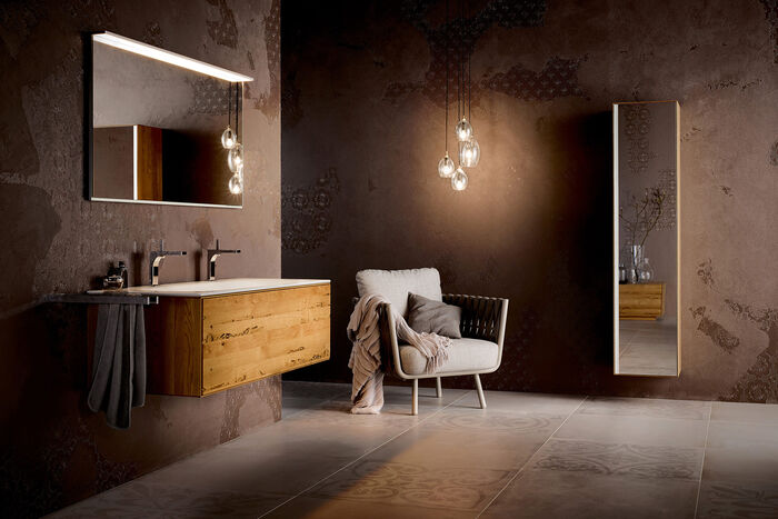 In Braun gehaltenes Badezimmer von KEUCO. Links befindet sich ein Waschbecken mit Unterschrank aus Holz und Spiegel. In der Mitte steht ein Sessel, über dem ein Designlicht hängt. An der Wand rechts hängt ein Ganzkörperspiegel.