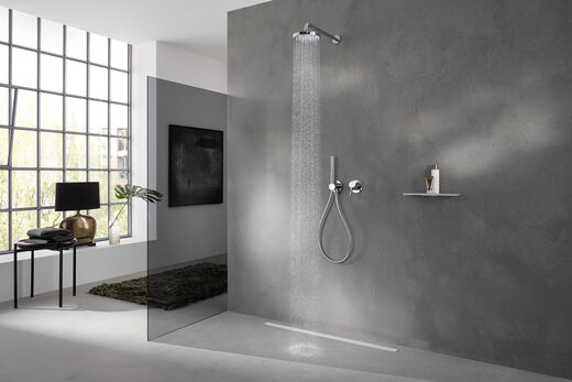 Helles, in grau-weiß gehaltenes Badezimmer, in dessen Dusche eine Keuco Ixmo Armatur installiert ist. Aus der Kopfbrause fließt Wasser, auf einer Ablagefläche stehen Duschutensilien. Links befindet sich ein Fenster, davor steht eine Stuhl, auf dem Handtücher liegen.