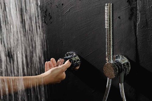 Keuco Ixmo Duscharmatur, die gerade von einer weiblichen Hand betätigt wird. Die Armatur ist an einer rustikalen schwarzen Wand befestigt, links strömt ein Wasserstrahl herab.
