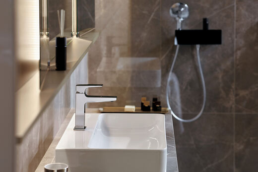 Eckiges Waschbecken mit Metropol - Armatur von hansgrohe. Links ist ein beleuchteter Spiegelschrank zu erkennen, im Hintergrund befindet sich eine begehbare Dusche.