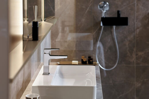 hansgrohe Metropole Eckiges Waschbecken mit Metropol - Armatur von hansgrohe. Links ist ein beleuchteter Spiegelschrank zu erkennen, im Hintergrund befindet sich eine begehbare Dusche.