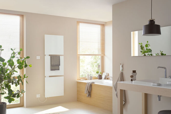 Elektro Desing Badheizkörper weiß eingeteilt in 3 Quadrate an der Wand montiert neben einer Badewanne und seitlich ein Waschplatz. 