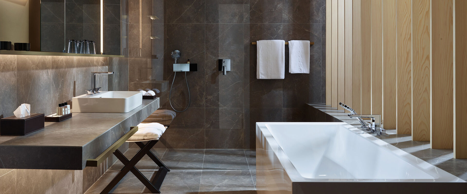 Modernes Badezimmer mit großer Badewanne, Waschbecken mit beleuchtetem Spiegelschrank, begehbarer Dusche und Ablagemöglichkeiten. Badarmaturen Metropol von Hansgrohe setzen Designakzente.