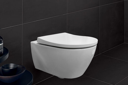 Designbetont und schlank: der WC-Sitz Slim von Geberit. In trendigen Wrap-over-Design schließt der Deckel bündig mit der Keramik ab, der eigentliche Sitz bleibt unsichtbar. Weißes WC vor schwarzen Fliesen.