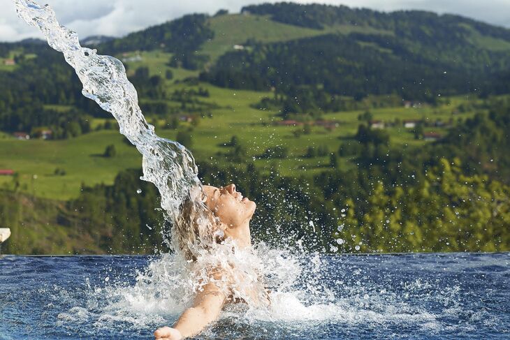 Wasserschwall trifft auf eine Frau im aussenpool vor einer Berglandschaft.