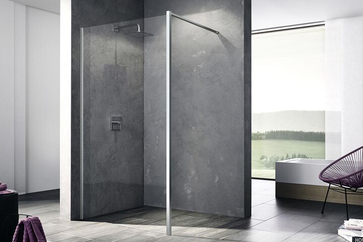 Bodenebene Dusche, an der eine Kermi Walk In XB Wall installiert ist. Die Duschwand aus Glas trennt die Dusche vom restlichen Badezimmerraum, dessen Boden gefliest ist. Rechts befindet sich eine Badewanne und ein Stuhl.