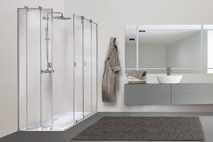 Helles Badezimmer, in dem links eine Duscholux-Pulsar Dusche steht, rechts daneben hängt ein Bademantel. Auf dem Boden liegt ein Teppich, rechts ist ein Waschbecken mit Unterschrank und Spiegeln installiert.