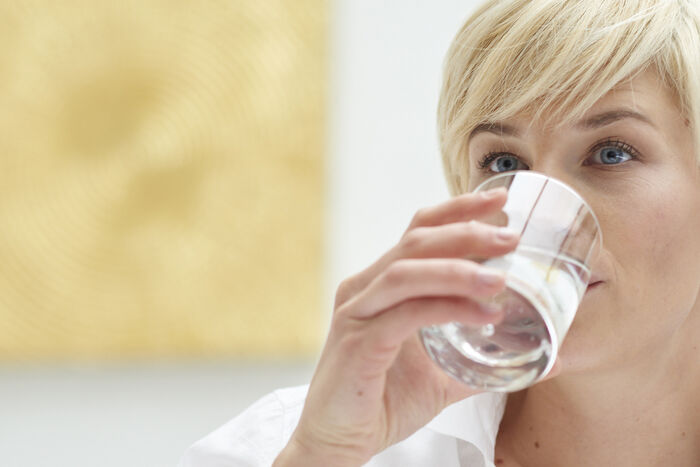 Frau mit Wasserglas aus dem sie trinkt im Hintergrund ein goldenes Bild.